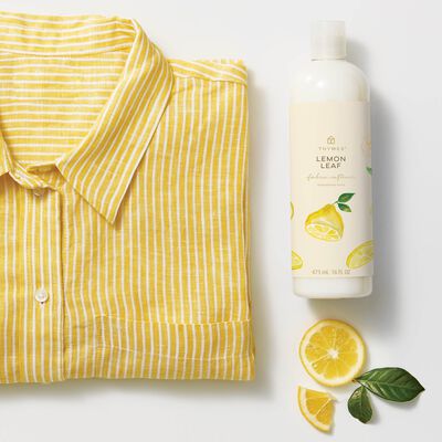Thymes Lemon Leaf Fabric Softener to Soften Clothing with Citrus Scent next to yellow shirt and lemon slices